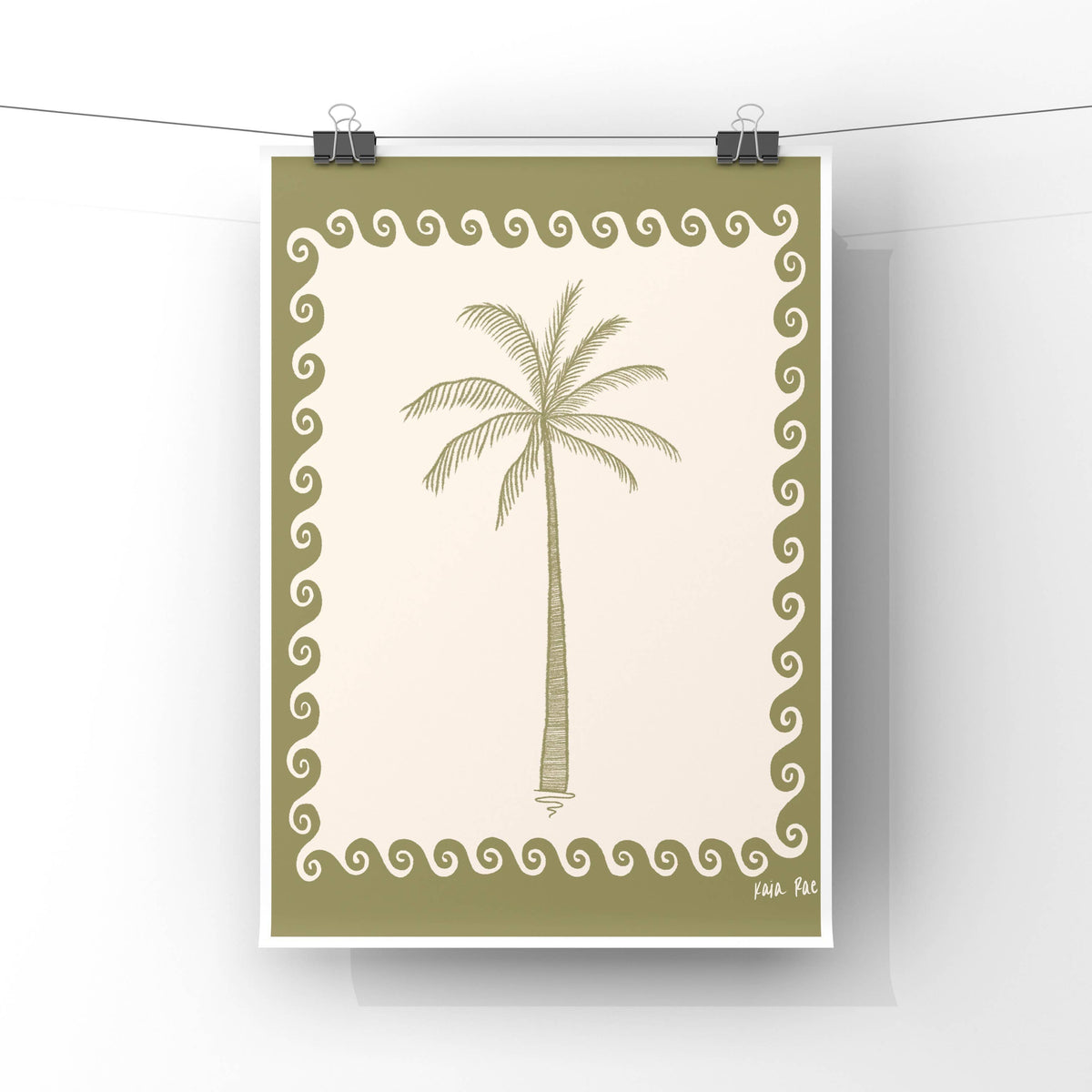 A vertical art print featuring a palm tree illustration, hanging from clips on a wire against a white wall. The print has an olive green border with a repeating scroll pattern and a cream center background.