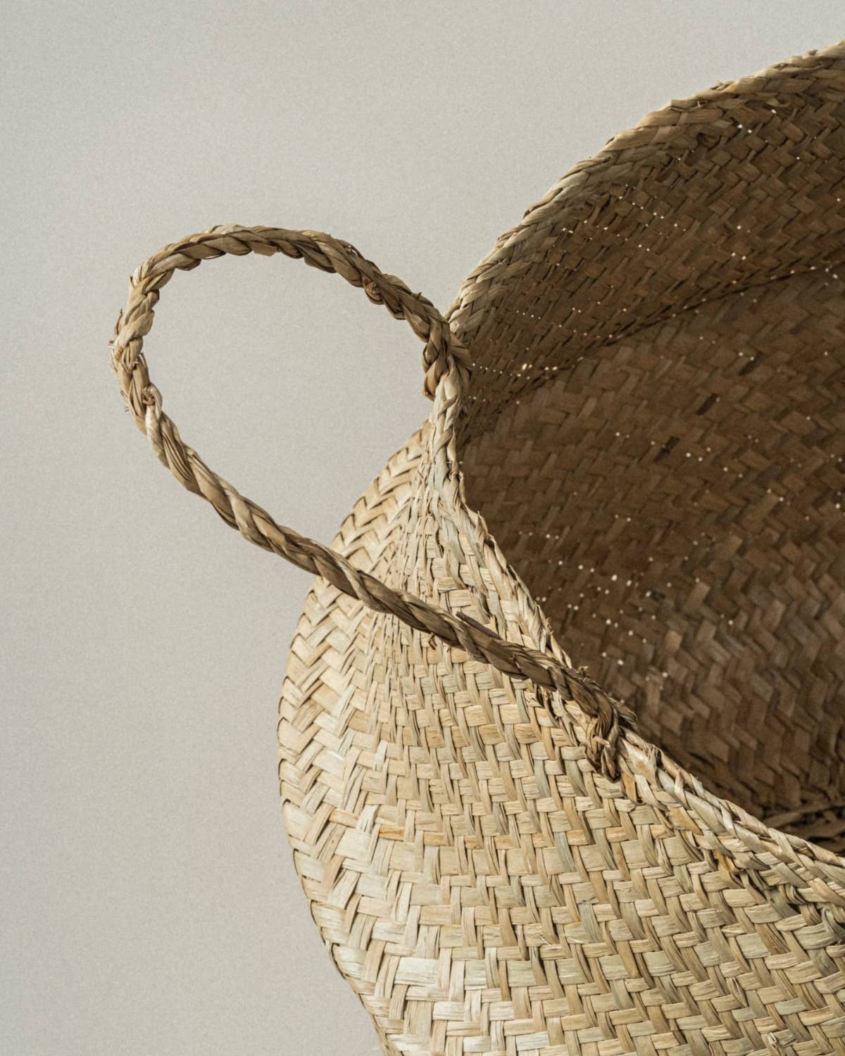 Close-up of a woven straw basket with a single braided handle, showing the natural beige and brown texture of the material against a plain light gray background.