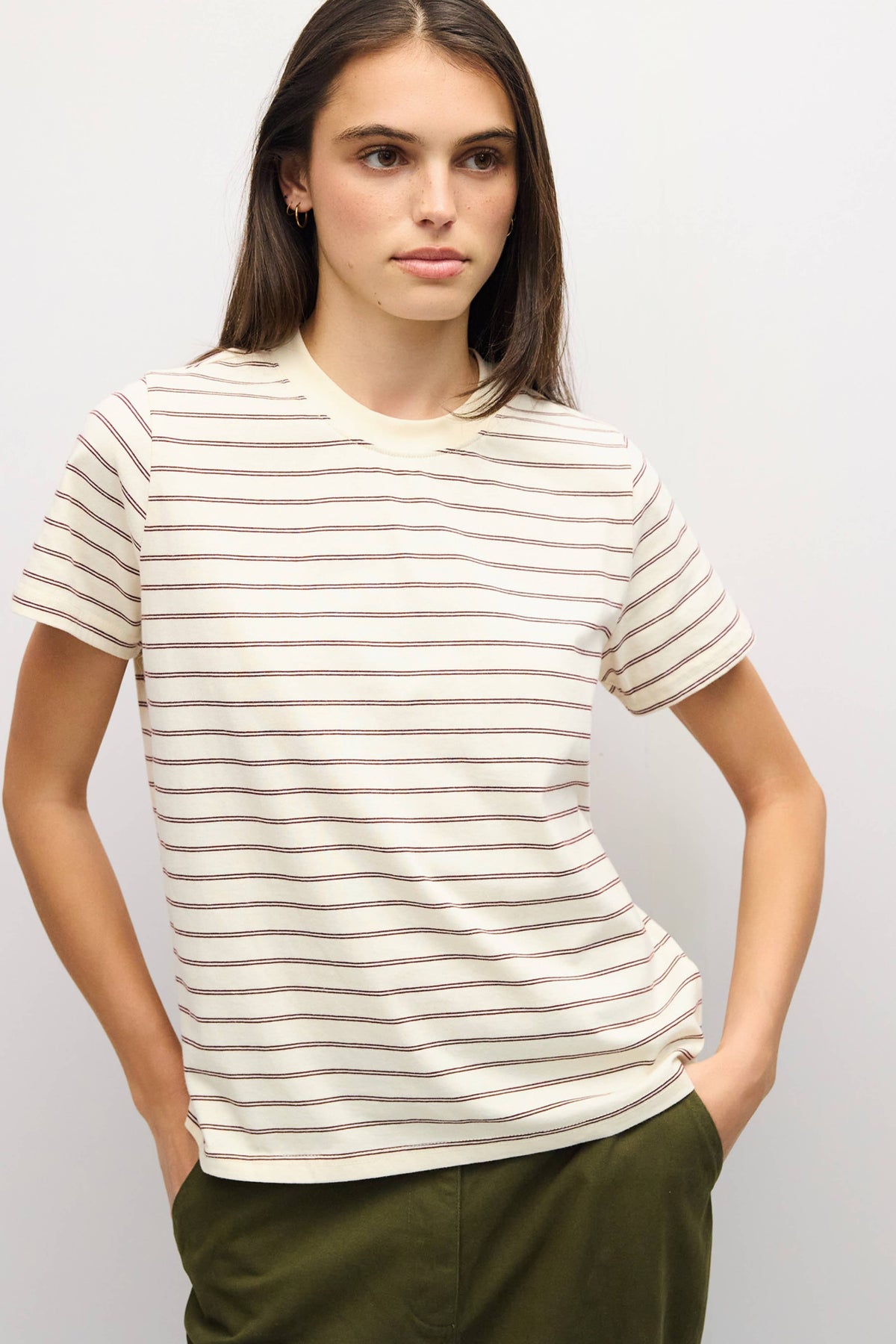 A woman wearing a short-sleeve crewneck t-shirt with thin horizontal beige stripes on an off-white base, paired with olive green pants, against a light gray studio background.