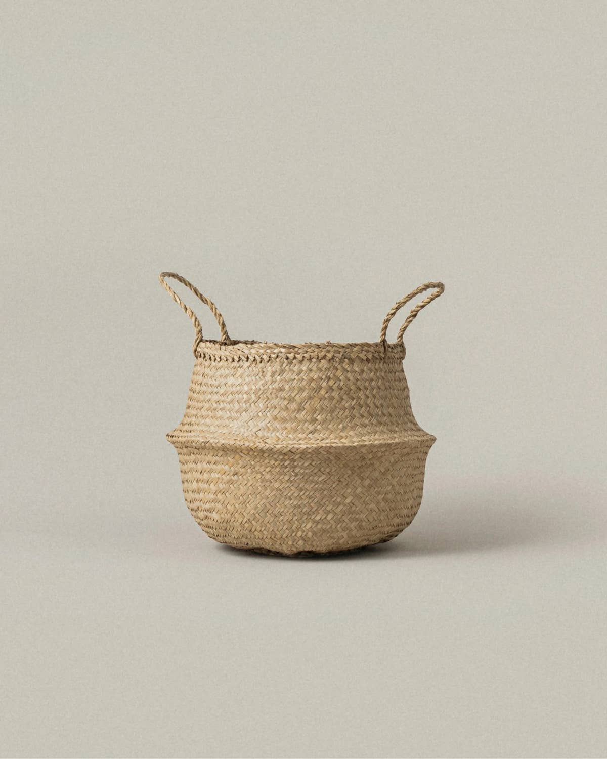 A natural woven basket made of straw or seagrass with two loop handles, shown on a plain light gray background.