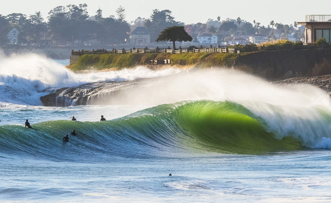 Surfers and a large wave in the ocean with a Sant Cruz background.