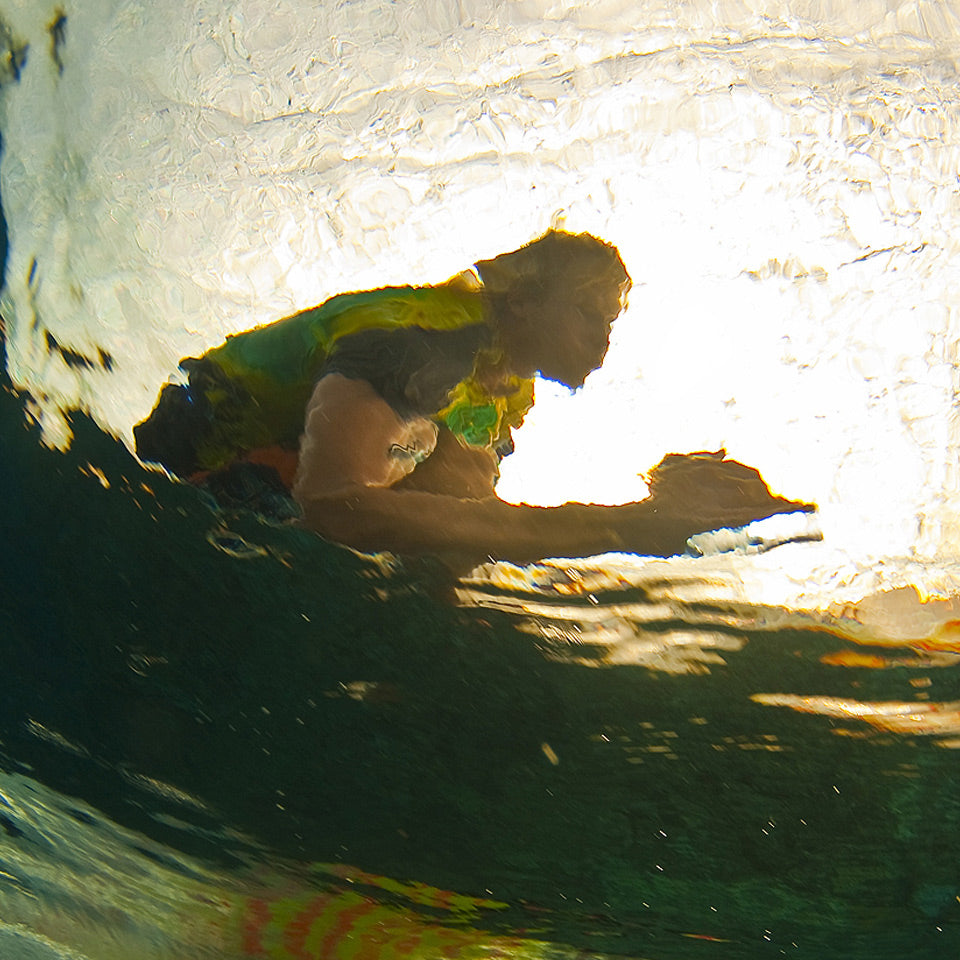 A person swimming underwater in a bright, sunlit pool, wearing a green and yellow rash guard or life vest over a dark swimsuit.