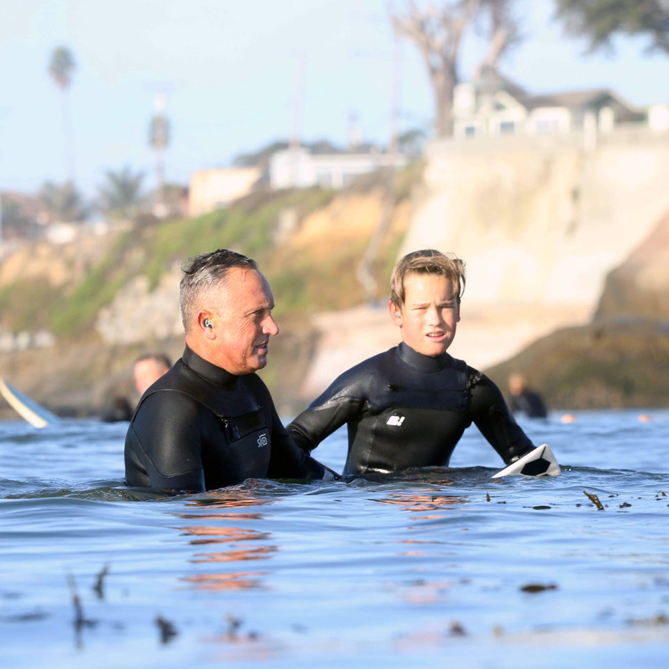 Two men in black wetsuits standing in ocean water near a beach, with a surfboard partially visible on the left and a rocky shoreline with houses in the background.